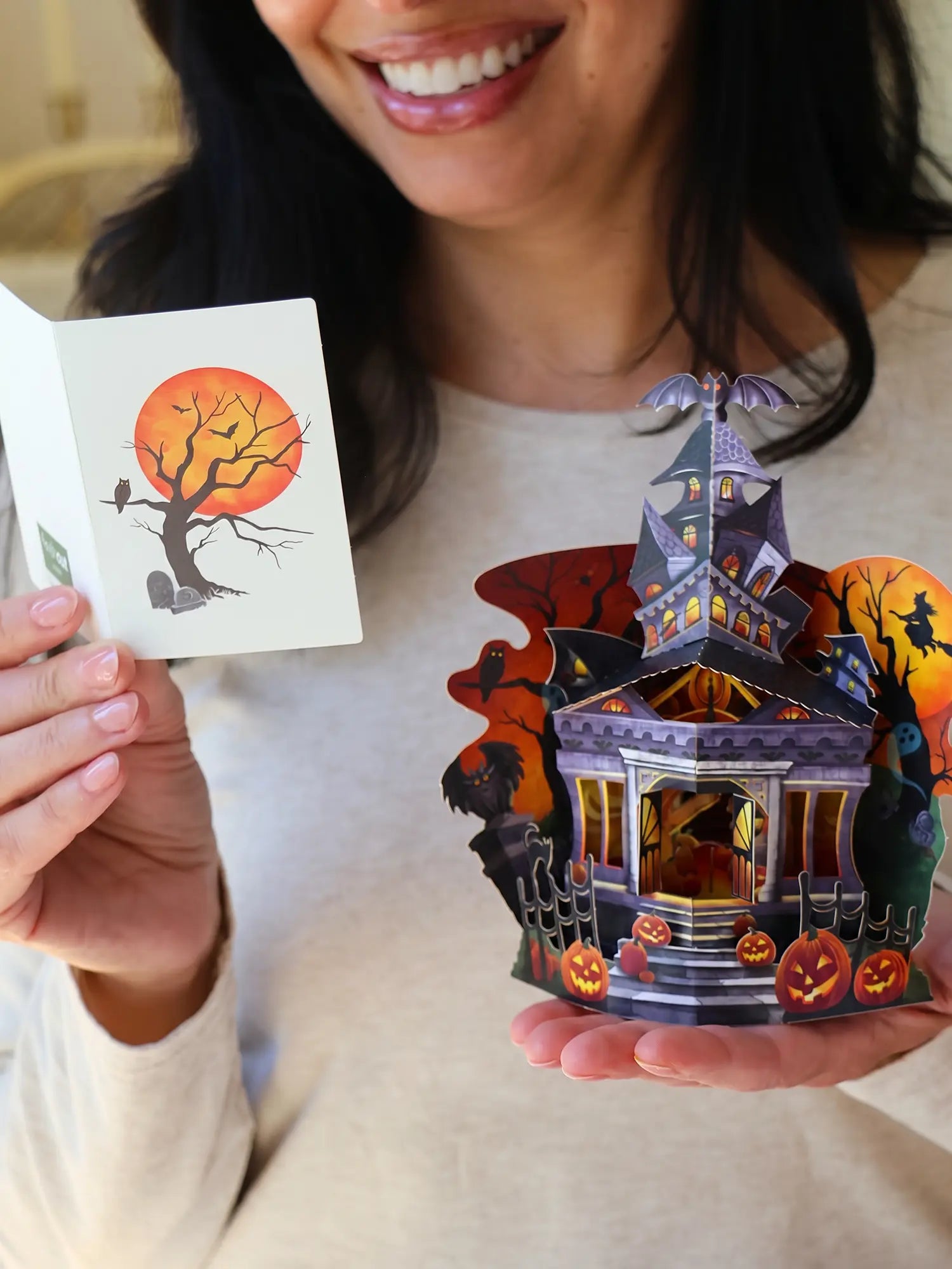 Person holding a pop-up card with a haunted house design and a matching greeting card featuring a spooky tree in front of a glowing orange moon.