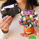 A woman holds a paper Halloween black cat in one hand and a colorful 3D paper flower bouquet in a jack-o'-lantern pot in the other. The bouquet features small white ghosts, a purple 'Happy Halloween!' banner, and a green 'Glow in the Dark' badge overlay.