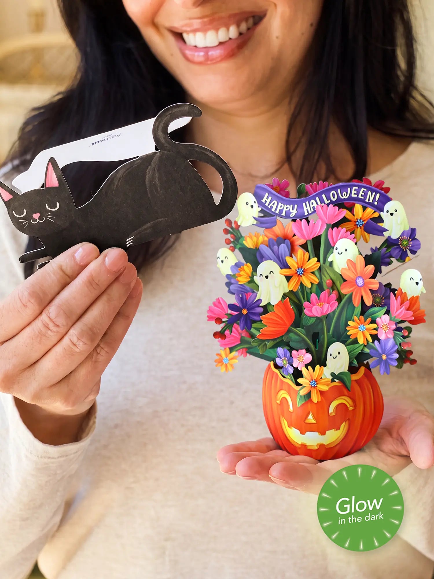 A woman holds a paper Halloween black cat in one hand and a colorful 3D paper flower bouquet in a jack-o'-lantern pot in the other. The bouquet features small white ghosts, a purple 'Happy Halloween!' banner, and a green 'Glow in the Dark' badge overlay.
