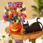 Halloween floral arrangement in a carved orange pumpkin pot with colorful flowers, ghost accents, and a purple 'Happy Halloween!' banner, displayed on a wooden stand beside a paper black cat with white pumpkins in the foreground.