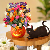 Halloween floral arrangement in a carved orange pumpkin pot with colorful flowers, ghost accents, and a purple 'Happy Halloween!' banner, displayed on a wooden stand beside a paper black cat with white pumpkins in the foreground.