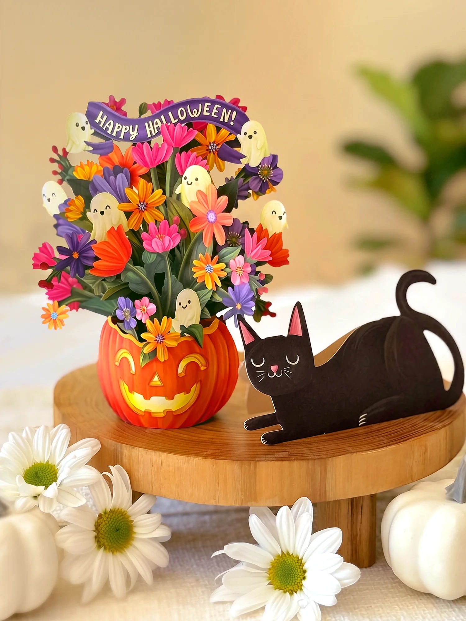 Halloween floral arrangement in a carved orange pumpkin pot with colorful flowers, ghost accents, and a purple 'Happy Halloween!' banner, displayed on a wooden stand beside a paper black cat with white pumpkins in the foreground.