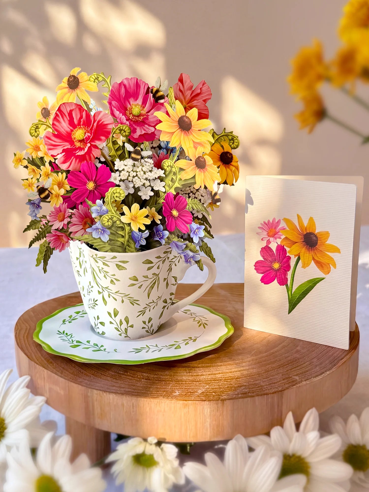 A colorful flower bouquet arranged in a white floral teacup and saucer, displayed on a round wooden stand beside a matching greeting card with flower illustrations.