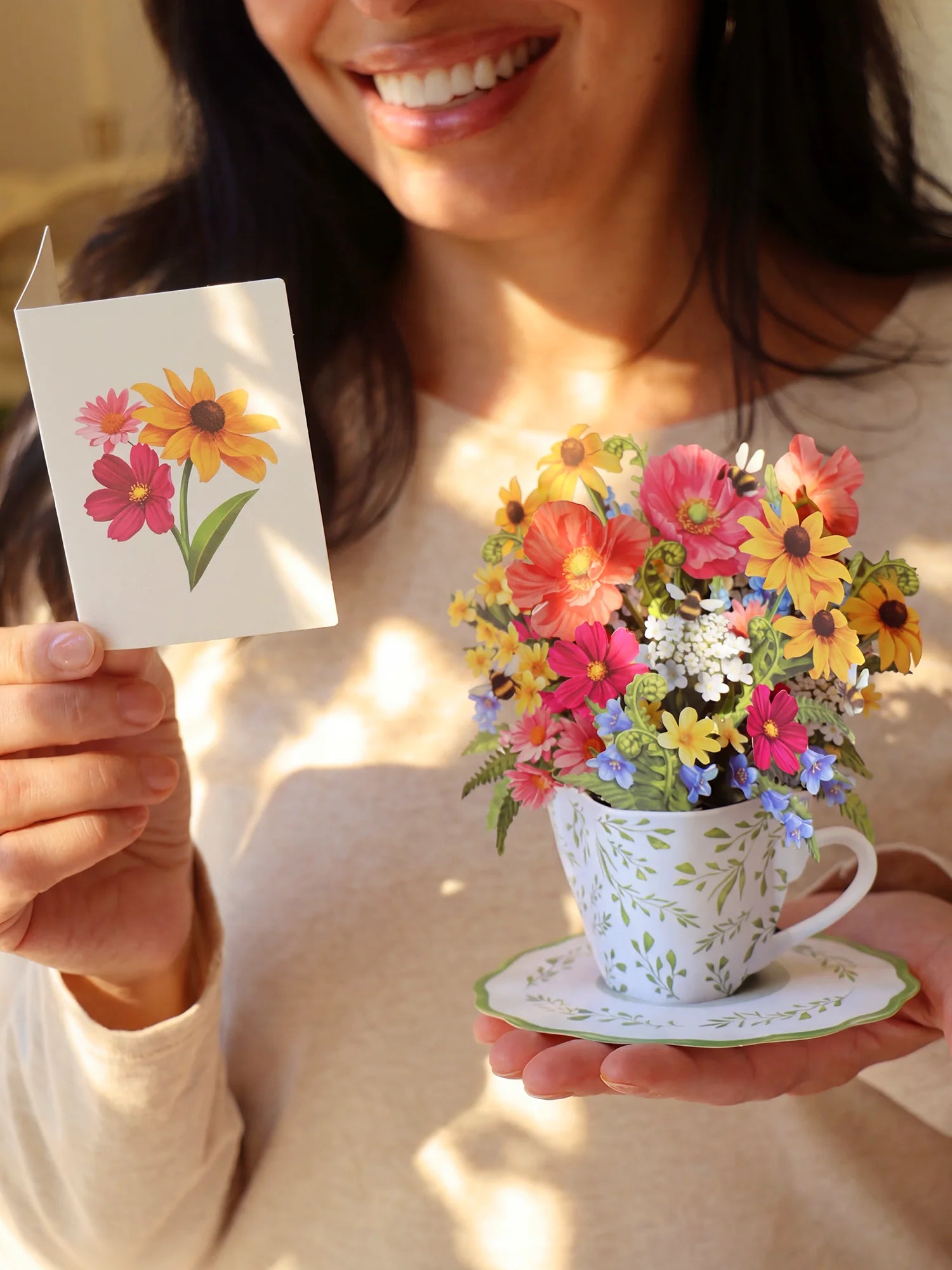 A smiling woman holds a small greeting card in one hand and a decorative teacup filled with colorful artificial flowers in the other, set against a softly lit indoor background.