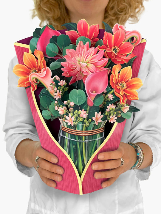 FreshCut Paper Dear Dahlia, is a symbol of kindness, is the centerpiece of this colorful pink-hued bouquet. Accented with the Cala Lily’s wishes of hope and rebirth, intertwined with smokey blue eucalyptus branches, this darling bouquet delivers a smile that will endure as long as the never-wilt blossoms. Our pop up 3D greeting cards are perfect for any occasion.
