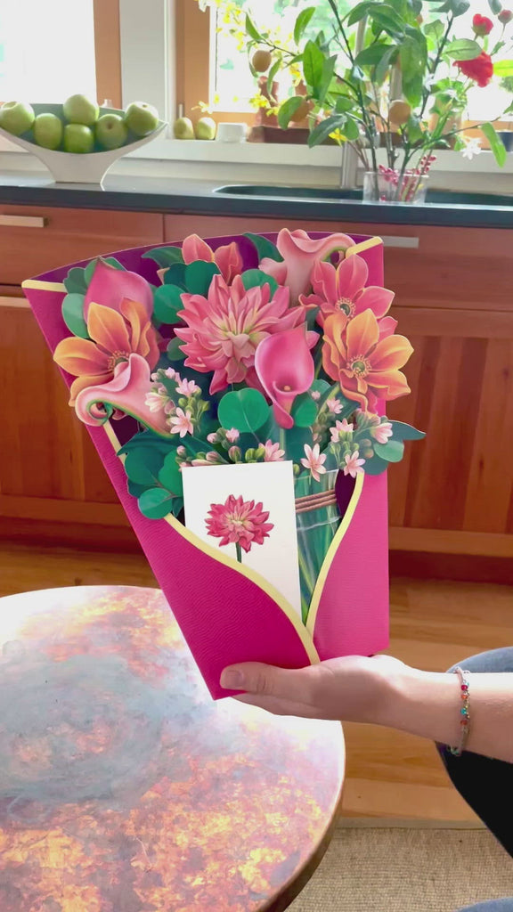Watch our FreshCut Paper Dear Dahlias bloom!, Dear Dahlia is a symbol of kindness, is the centerpiece of this colorful pink-hued bouquet. Accented with the Cala Lily’s wishes of hope and rebirth, intertwined with smokey blue eucalyptus branches, this darling bouquet delivers a smile that will endure as long as the never-wilt blossoms. Our pop up 3D greeting cards are perfect for any occasion.